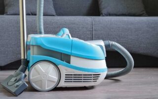 A blue vacuum cleaner on a wooden floor near a sofa highlights effective methods for cleaning laminate flooring. Keep your floors spotless and durable with expert tips from In Home Flooring, your trusted Denver flooring expert. Discover the best tools and techniques to maintain the beauty of your laminate floors.