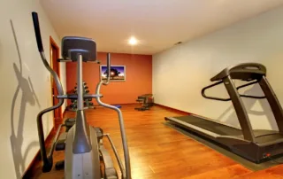 Flooring for home gyms at high altitude focuses on subfloor deflection and impact ratings, illustrated by a gym setup with a treadmill and elliptical. The scene shows how proper floor stability, cushioning, and support help equipment perform safely in demanding conditions.