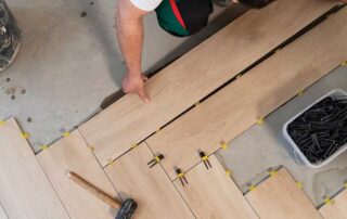 Person installing wooden floor tiles, answering the question: Should I start with cabinets or flooring for an efficient and seamless home renovation?
